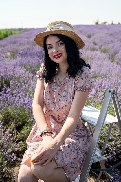 Giovane bruna con un cappello di paglia si siede su uno sfondo di lavanda in un abito bianco a pois