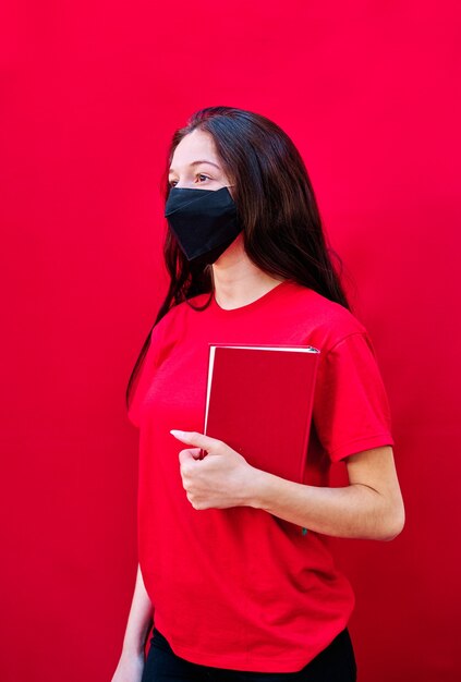Молодая брюнетка студентка с маской, несущая книгу на плоском красном фоне