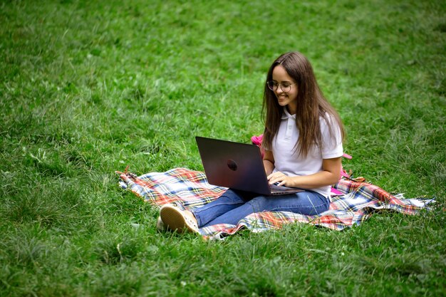 공원 잔디밭에 깔개에 앉아있는 동안 노트북으로 작업하는 젊은 갈색 머리 학생