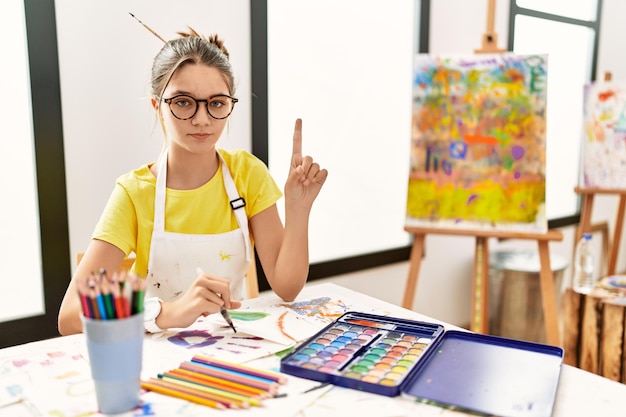 Young brunette teenager at art studio pointing with finger up and angry expression showing no gesture