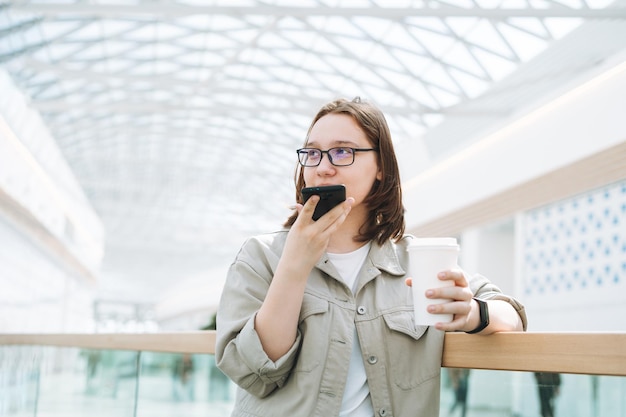 휴대전화를 사용하여 안경을 쓴 젊은 브루네트 10대 여학생이 공공 장소 쇼핑몰에서 종이 커피 컵과 함께 음성 메시지를 보냈습니다.
