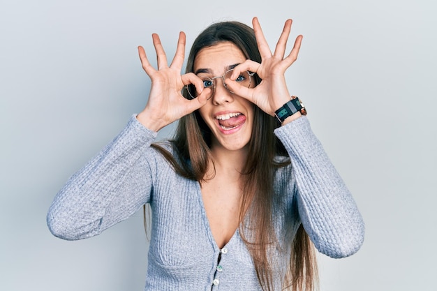 young brunette teenager wearing casual sweater and glasses doing ok gesture like binoculars sticking tongue out eyes looking through fingers crazy expression