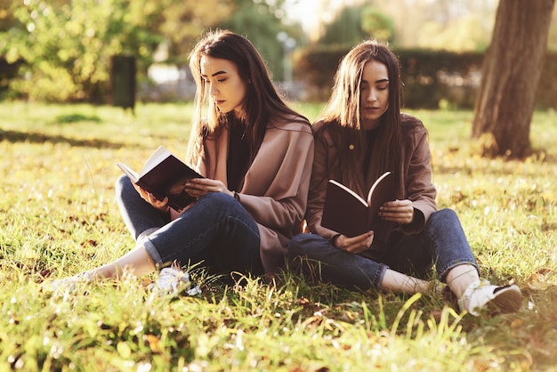 Giovani sorelle gemelle brunetta seduti vicini l'uno all'altro sull'erba con le gambe leggermente piegate in ginocchio e incrociate, leggendo libri marroni, indossando un cappotto casual nel parco soleggiato di autunno su sfondo sfocato.