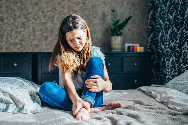 Young brunette in a white blouse and blue jeans sits on a bed in her room, clutching a sore leg