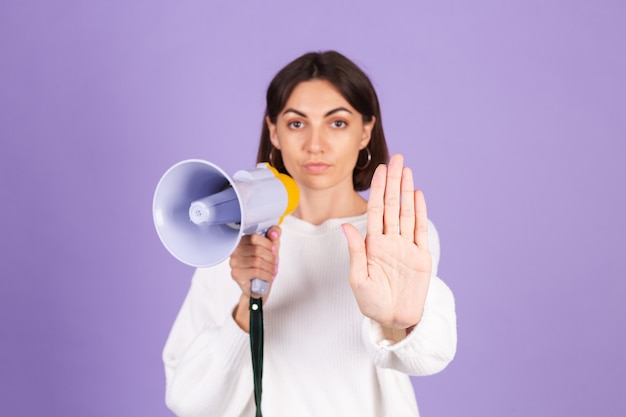 Young brunette in white casual sweater isolated on purple wall with megaphone doing stop sing with palm of the hand