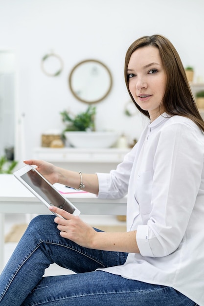 A young brunette in a white shirt and jeans writes SMS on the phone A girl sits on the bed and reads the message text on the smartphone Hotel room Homeliness