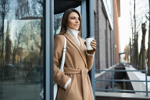 Giovane bruna con una tazza di caffè vicino all'ingresso dell'edificio aziendale