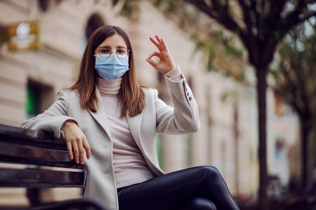 Young brunette with face mask sitting on the bench outside and showing okay gesture. Millennials during corona outbreak.