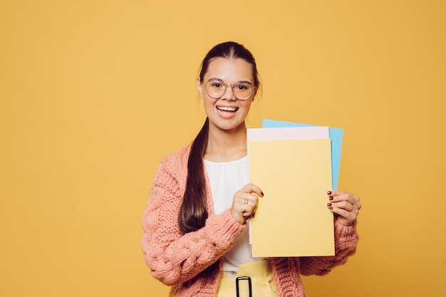 Young brunette with long hair dressed in a white blouse, pink sweater and yellow pants holding a sheet of colored paper in front of her.Cute smiling standing on a yellow background with a copy space.