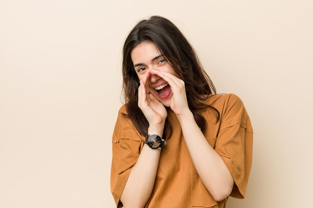 Young brunette woman against a beige background shouting excited to front.