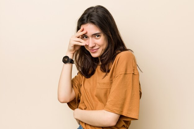 Young brunette woman against a beige blink at the camera through fingers