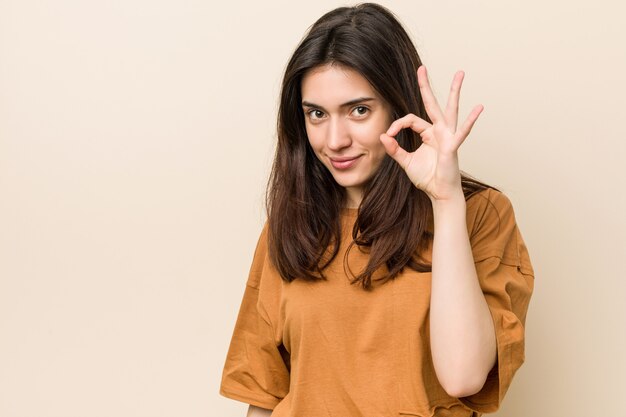 Young brunette woman against a beige wall cheerful and confident showing ok gesture.