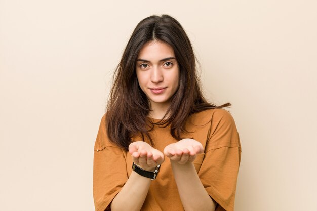 Young brunette woman against a beige wall holding something with palms, offering