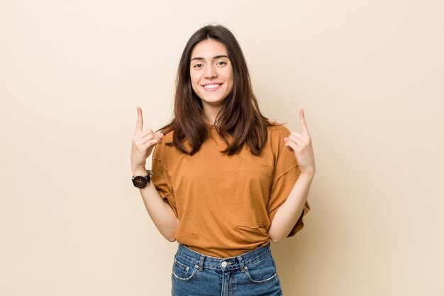 Young brunette woman against a beige wall indicates with both fore fingers up showing a blank space.