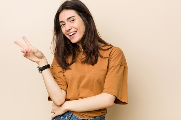Young brunette woman against a beige wall joyful and carefree showing a peace symbol with fingers.
