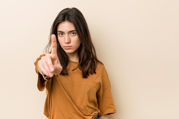 Young brunette woman against a beige wall showing number one with finger.