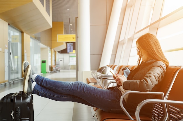 Young brunette woman in airport is waiting for her flight something looking at smartphone