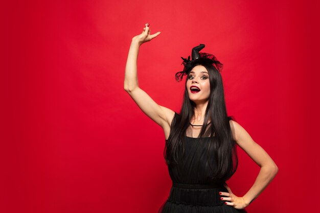 Young brunette woman in black hat and costume on red background. Attractive caucasian female model. Halloween, black friday, cyber monday, sales, autumn concept. Copyspace. Pointing up.