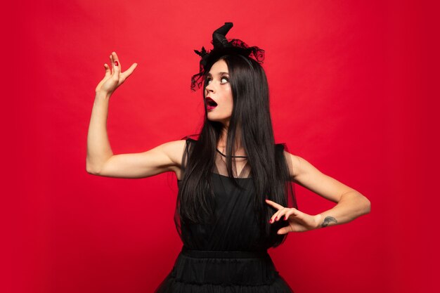 Young brunette woman in black hat and costume on red background. Attractive caucasian female model. Halloween, black friday, cyber monday, sales, autumn concept. Copyspace. Pointing up.