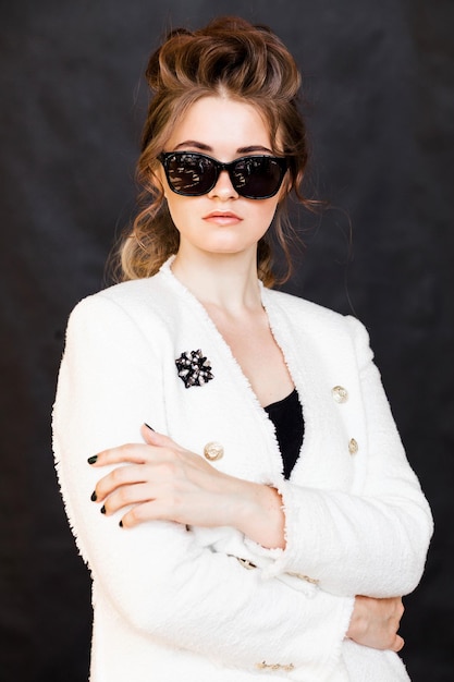 Young brunette woman in black sunglasses and a white jacket with a brooch posing on a black background