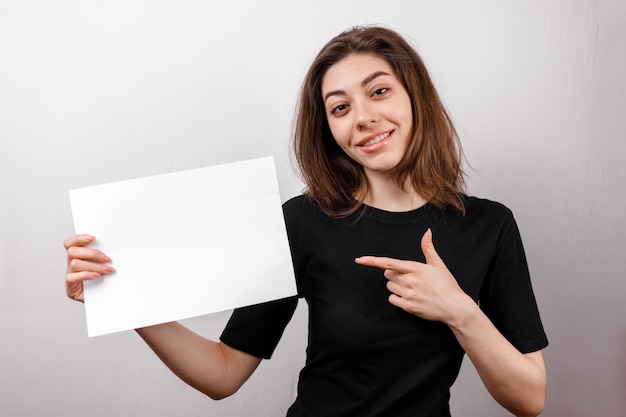 Young brunette woman in a black t-shirt smiling shows a white sheet on white