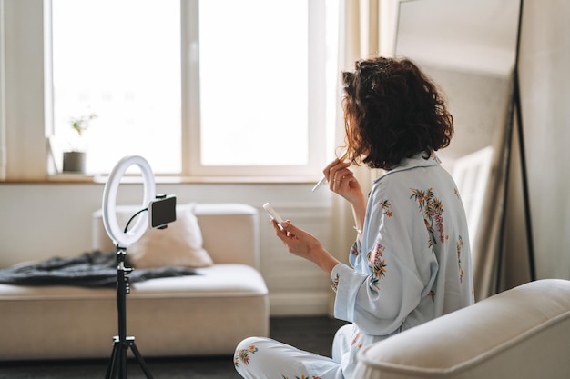 young brunette woman blogger in home clothes doing makeup with mobile phone and ring lamp online