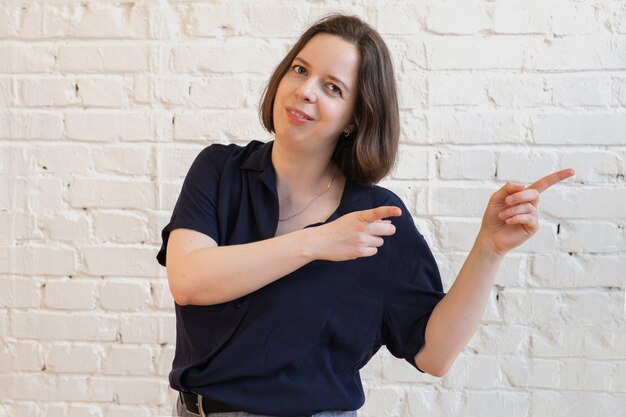 Young brunette woman in a blue blouse on white brick wall background. The woman smiles and points her fingers to the right.