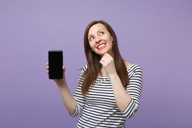 Young brunette woman in casual striped clothes posing isolated on violet purple background studio portrait. People lifestyle concept. Mock up copy space. Hold in hand cellphone with blank empty screen