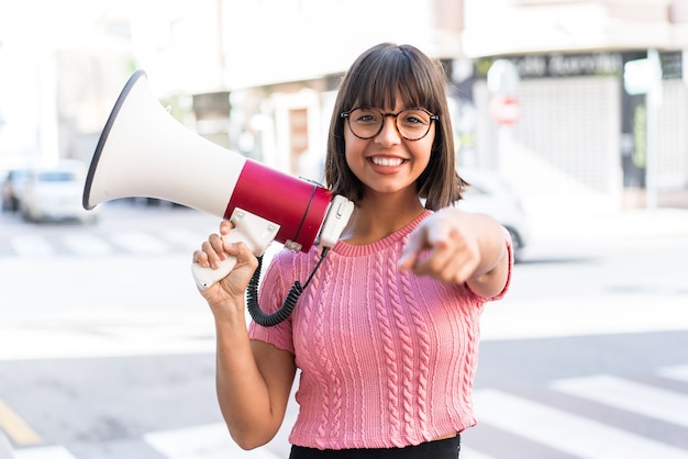 Young brunette woman in the city holding a megaphone and smiling while pointing to the front