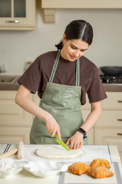 La giovane donna castana cuoca in grembiule verde taglia la pasta stesa con un coltello di plastica su una teglia in silicone