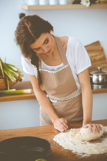 Young brunette woman cooking pizza or handmade pasta in the kitchen. Housewife preparing dough on wooden table. Dieting, food and health concept.