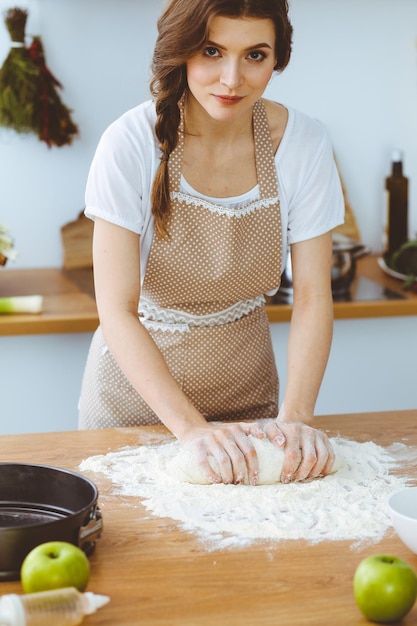 Young brunette woman cooking pizza or handmade pasta in the kitchen. Housewife preparing dough on wooden table. Dieting, food and health concept.