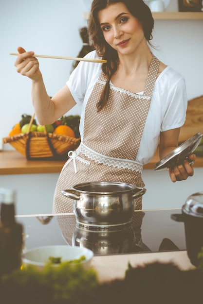 Young brunette woman cooking soup in kitchen. Housewife holding wooden spoon in her hand. Food and health concept.