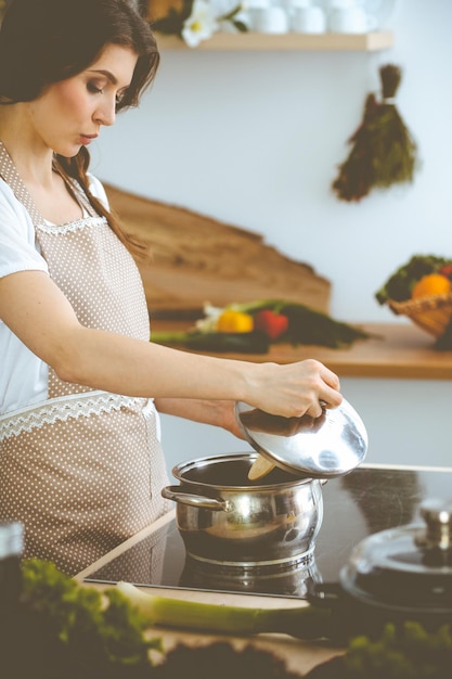 Young brunette woman cooking soup in kitchen. Housewife holding wooden spoon in her hand. Food and health concept.