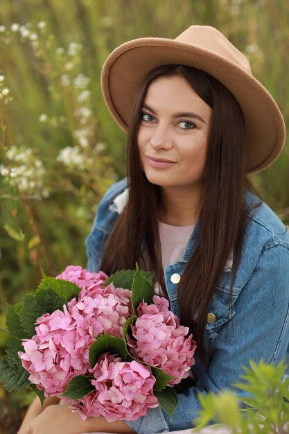 Young brunette woman in denim jacket, pink dress and hat holding bouquet of pink flowers hydrangea sitting in field on summer day.