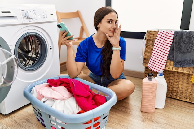 Young brunette woman doing laundry using smartphone hand on mouth telling secret rumor whispering malicious talk conversation