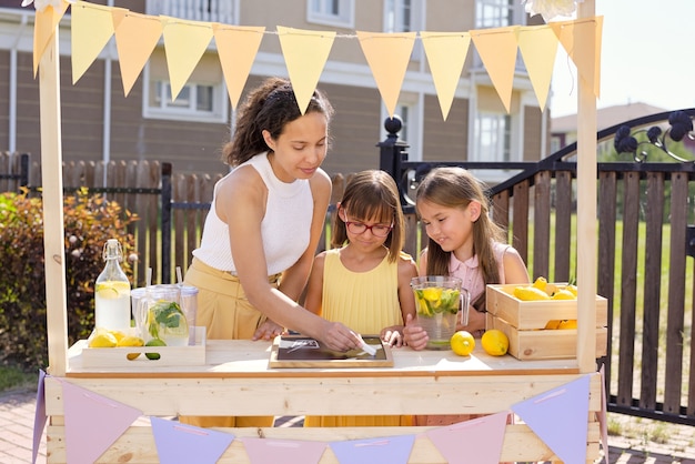 Young brunette woman drawing glass of drink on noticeboard with chalk while her daughters standing near by and selling lemonade by stall