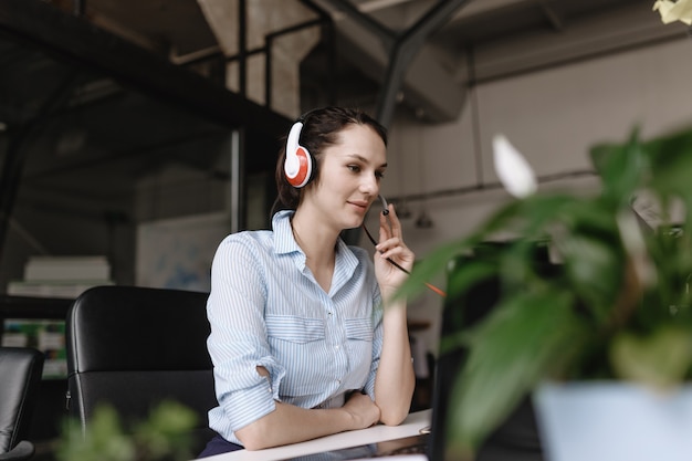 Young brunette woman dressed in office style clothes is talking to customers through the headset sitting at desk with laptop in the modern office