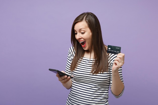 Young brunette woman girl in casual striped clothes posing isolated on violet purple background studio portrait. people lifestyle concept. mock up copy space. hold in hand credit bank card cellphone