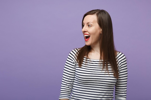 Young brunette woman girl in casual striped clothes posing isolated on violet purple wall background studio portrait. People sincere emotions lifestyle concept. Mock up copy space Looking aside smile.