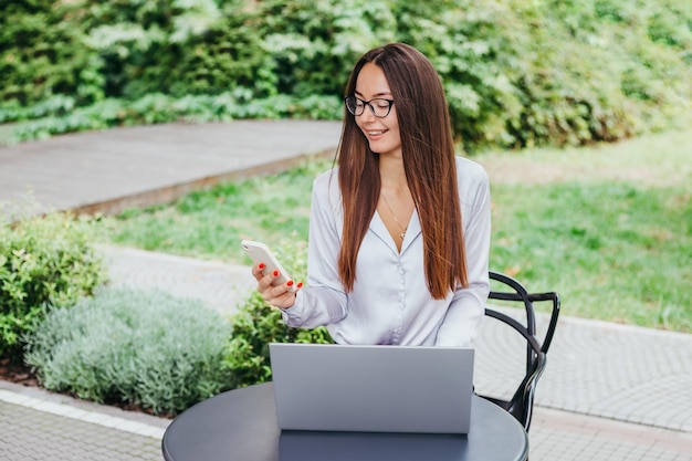 A young brunette woman in glasses sits at a table in a cafe and works at a laptop