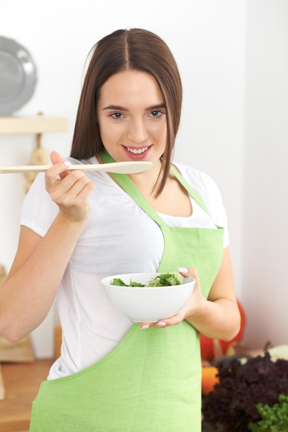 Giovane donna bruna in grembiule verde sta cucinando o mangiando insalata fresca in cucina. casalinga che tiene cucchiaio e ciotola di legno nelle sue mani. concetto di cibo e salute.