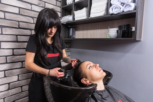 Young brunette woman having her hair washed by female professional stylist in washing sink in beauty salon