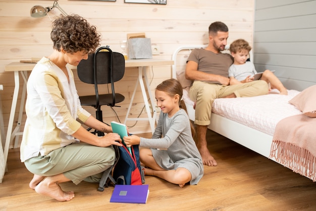 Young brunette woman helping her little daughter with packing school rucksack with copybooks and other stuff on the floor of bedroom
