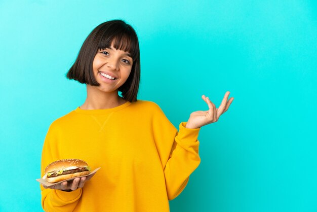 Young brunette woman holding a burger over isolated background extending hands to the side for inviting to come