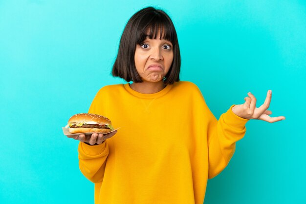 Foto giovane donna castana che tiene un hamburger sopra fondo isolato che ha dubbi mentre alza le mani