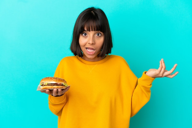 Photo young brunette woman holding a burger over isolated background with shocked facial expression