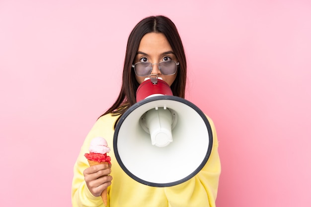 Young brunette woman holding a cornet ice cream on isolated pink shouting through a megaphone