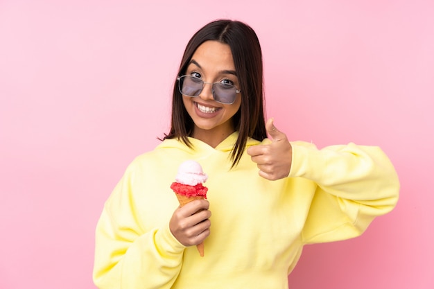 Giovane donna castana che tiene un gelato della cornetta sopra la parete rosa isolata che mostra segno e pollice giusti sul gesto