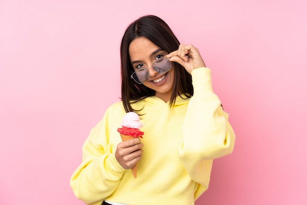 Young brunette woman holding a cornet ice cream over isolated pink wall with glasses and happy